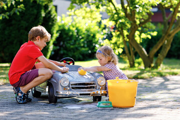 Two happy children washing big old toy car in summer garden, outdoors. Brother boy and little...