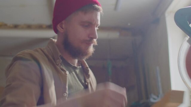 Tilt up shot of young bearded carpenter in red hat cutting wooden plank with miter saw while working in workshop