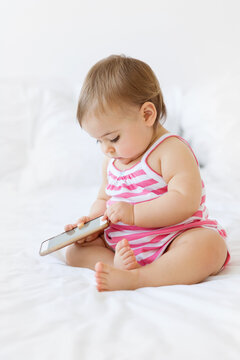 Chubby Baby Girl Sitting On A White Bed Holding Smartphone Pressing Button With Finger