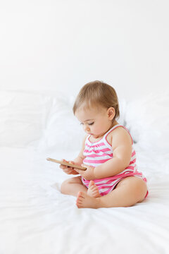 chubby baby girl sitting on a white bed holding smartphone looking down at screen