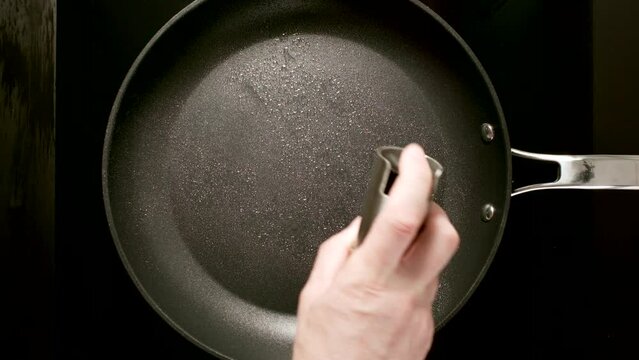 TOP VIEW: Man's hand sprays vegetable oil from dispenser (spray gun) on a black frying pan on stove