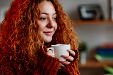 A cute ginger girl with curly hair is sitting in the chair at home in the morning and drinking her coffee.