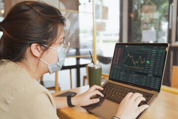 Asian Businesswoman, female trader, investor with medical face mask using laptop, tablet for stock market, Bitcoin cryptocurrency trading, money investing on online trading platform. Trading concept.