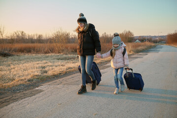 Mother and daughter are walking along the road and carrying suitcases on wheels. Refugees.