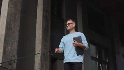 A young man is going down a stairs business center. He is holding a laptop. He is smiling. He is wearing glasses and a sweater. 4k