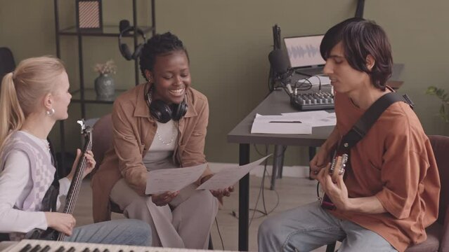 Medium slowmo of young multiethnic music band playing music instruments and singing while rehearsing for concert at home studio