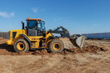 Fototapeta premium Bulldozer or loader moves the earth at the construction site against the blue sky. An earthmoving machine is leveling the site. Construction heavy equipment for earthworks.