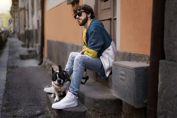 a young hipster with sunglasses is resting on the steps in front of a house with his French bulldog - dog and owner through the streets of the old city