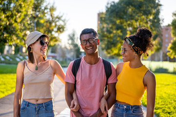 Happy people of generation z walking in a city park, handsome young man laughing while two beautiful women looking at him, summer moment of urban travel and tourism