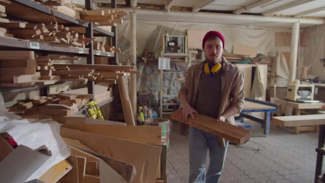 Artisan taking plank from shelf, then bringing it to workbench and putting on safety earmuffs before woodworking in carpentry workshop