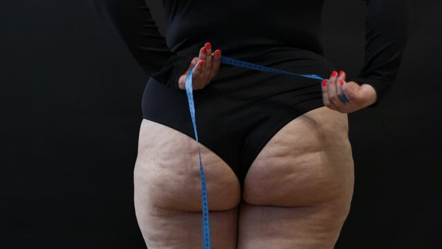 a fat woman with cellulite measures herself with a measuring tape on a black background