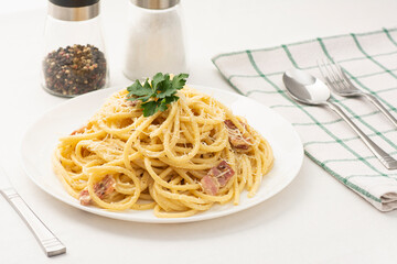 Cream and bacon Pasta with green parsley leaves on a white plate with a kitchen towel and salt and paper on background with fork, knife, and spoon. Italian spaghetti with pamezan.