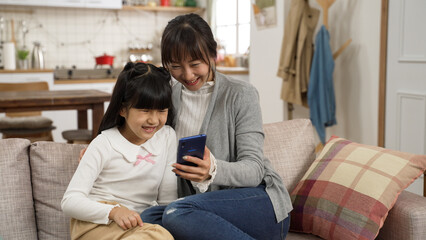 happy asian mother and daughter relaxing on couch watching funny online video with mobile phone. the girl touches the screen to play again
