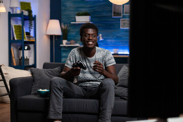 Happy excited african american gamer playing virtual video games using wireless joystick winning online gaming tournament. Winner player relaxing on couch in living room enjoying leisure time © DC Studio
