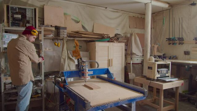 Wide shot of woodworker operating CNC machine with computer while drilling wooden plank in carpentry workshop