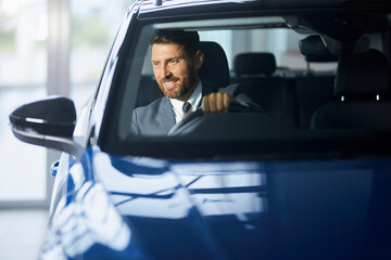 Positive bearded man in formal wear sitting on driver's seat of modern car and examining interior. Male customer choosing new transport at showroom.