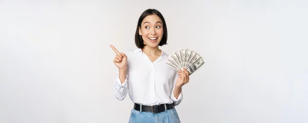 Fotobehang Smiling young modern asian woman, pointing at banner advertisement, holding cash money dollars, standing over white background © Mix and Match Studio