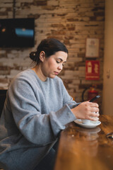 Chica joven guapa tomando té  en una cafetería muy acogedora con sudadera gris y mirando el smartphone