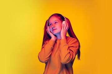 Portrait of young girl with two pontails listening to music in headphones isolated over yellow background in neon light