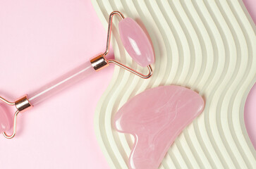 Rose quartz gouache massage scraper and facial massage roller on a beige wavy podium. Anti-aging...