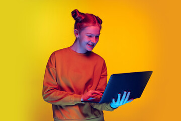 Portrait of excited young girl, looking on laptop, searching information isolated over yellow...