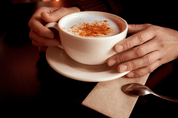 Female hands holding a cup of turkish traditional winter hot drink salep with cinnamon. White ceramic mug on plate and little spoon on napkin. Close-up.