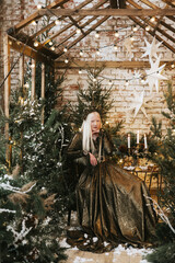 Fototapeta na wymiar young albino woman with blue eyes and long white hair in beautiful green dress and crown stands in loft room decorated with wooden greenhouse and Christmas trees with twinkle lights, diverse people