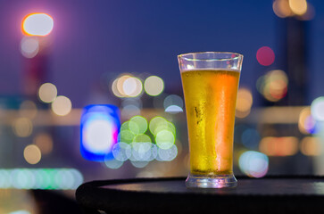 A glass of beer puts on table at rooftop bar with colorful city bokeh light background.