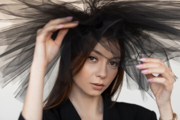 Fototapeta premium Young flawless woman with long dark hair, make-up wearing black blazer touching black organza decoration on head.