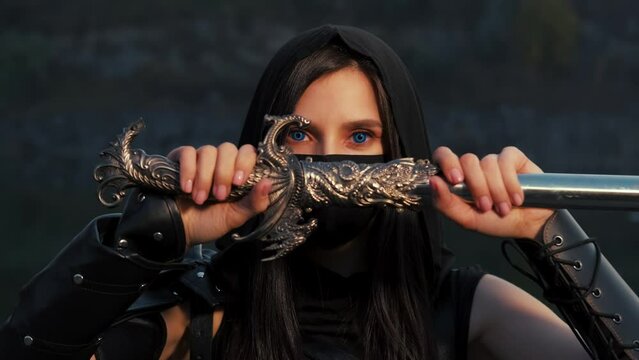close-up portrait fantasy woman warrior assassin holding dagger in hands, hiding face behind mask. Armed girl, blue eyes. Black leather costume hood on head. Ninja soldier with knife sword. 4k footage