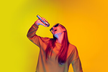 Portrait of young girl singing in microphone, posing isolated over yellow studio background in neon...