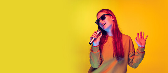 Portrait of young girl singing in microphone, posing isolated over yellow studio background in neon light. Flyer