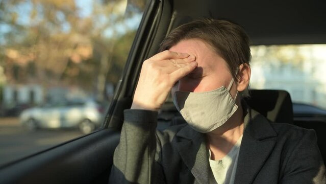 Businesswoman with severe headache pain symptoms and protective face mask at the backseat of a car