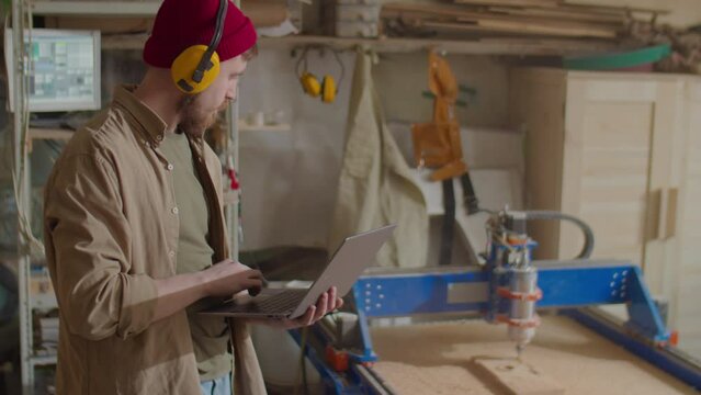 Carpenter in protective earmuffs using laptop while operating woodworking CNC machine in workshop