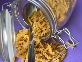 A tablespoon is in a glass jar with dry pasta. Lilac background.
