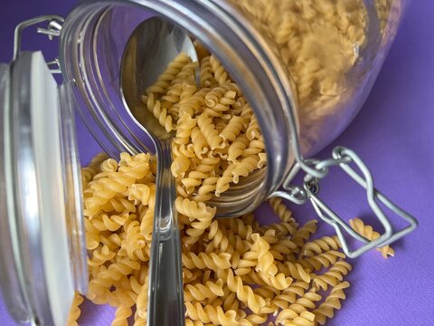 A tablespoon is in a glass jar with dry pasta. Lilac background.