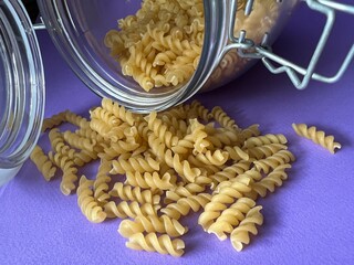 A glass jar with a scattering of dry twisted pasta. Lilac background.