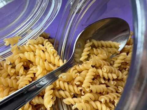 A tablespoon is in a glass jar with dry pasta. Lilac background.