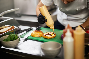 Crop view of chef's hands pouring sauce on cheeseburger in restaurant. Male hands in gloves...