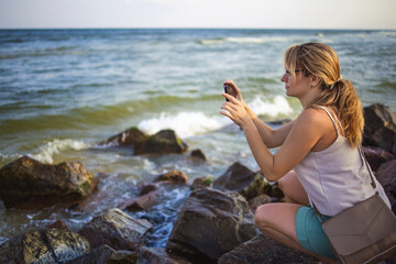 Woman photographs the sea landscape on the phone while sitting on the stones