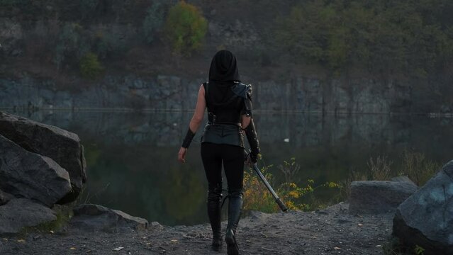 silhouette fantasy woman warrior assassin holds dagger in hands walks at night nature. Rear view, back armed girl. Black leather costume, hood. Ninja soldier with sword knife. 4k footage. river rocks.