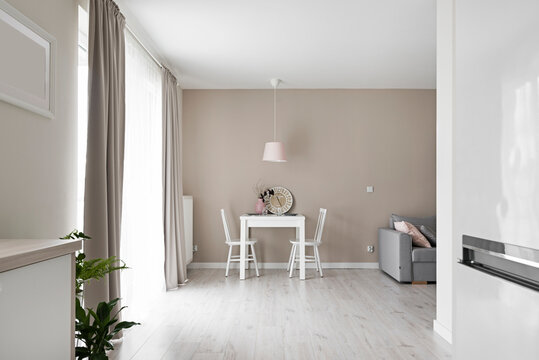 Salon i jadalnia w kolorach beżowych, białych i r&oacute;żowych. Firanka i zasłony. Szara sofa. Stolik do jedzenia z wisząca r&oacute;żową lampą.