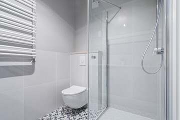 Nowoczesna jasna łazienka w kolorach białych i szarych. Toaleta, prysznic, blat z umywalka i kranem oraz grzejnik. 