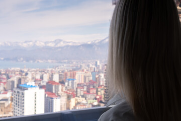 Fototapeta na wymiar Portrait of a blonde woman from the back looking from a height at a blurred city and mountains on a sunny day, copy space