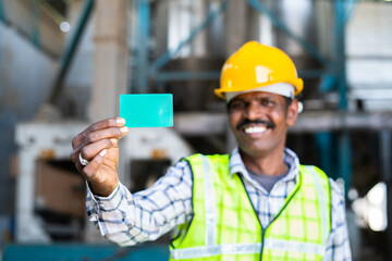 focus on hand, happy industrual worker holding green card by looking at camera in front of machinery at factory - concept employment or labour id.