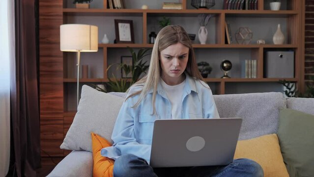 Young nervous woman sitting on sofa while working on laptop at home. She gets bad news, gets upset feels stress and hopelessness. Pretty girl freelancer having unexpected trouble feels tense.