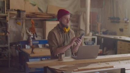 Young male woodworker with safety earmuffs over his neck using smartphone in carpentry workshop - Powered by Adobe