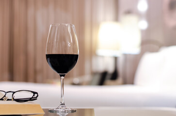 A glass of red wine with spectacles and book put on table in bed room. Relaxing at home concept.