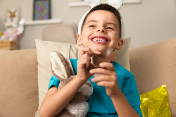 Joyful brazilian boy with easter bunny ears enjoying chocolate egg in the sofa