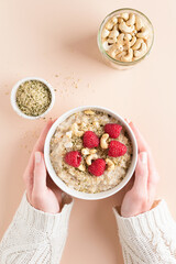 Bowl of oatmeal porridge with raspberries, hemp seeds and nuts in female hands over beige...